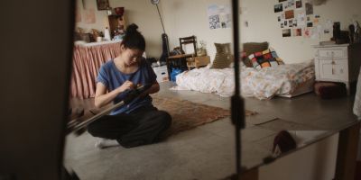 Film still from 'Sue's Apartment' (2024) by Jon Beaton. Reflected in a mirror, a young woman with dark hair sits cross-legged on a rug in a student apartment trying to fix a vacuum cleaner head.